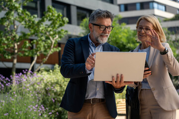 Diverse business couple collaborating on laptop outdoors in modern urban district, analyzing data and planning ideas together, teamwork.