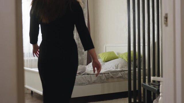 Back view teenager enters room confidently, turns to display black dress while friend seated on bed admires style, soft daylight filters through window, bed with green pillows visible