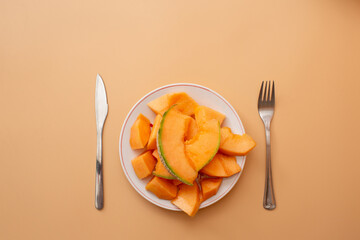 Closeup of sliced cantaloupe melon on a plate with fork and knife placed symmetrically on beige pastel background.