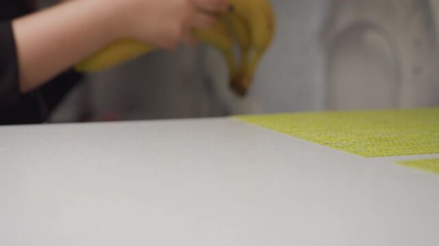 Close up manicured hands carefully lifting banana bunch from table into nylon bag, natural light highlights ripe fruit yellow peel, gesture emphasizes fresh produce, clean surface