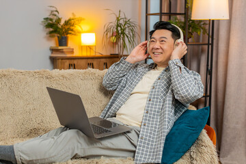 Happy relaxed overjoyed Chinese middle-aged man in wireless headphones sitting on home sofa choosing listening disco music while working on laptop netbook. Guy enjoys weekend activities entertaining.