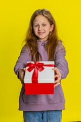 Fototapeta premium Lovely smiling young preteen child girl kid presenting birthday gift box stretches out hands, offer wrapped present career bonus, celebrating party. Children on studio yellow background. Vertical