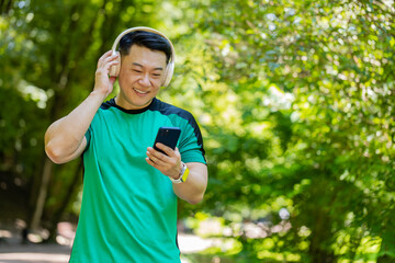 Athletic fitness adult guy sport runner man walking, wearing headphones listening music from smartphone and smiling. Healthy Japanese sportsman workout cardio outdoors in morning park on summer day.