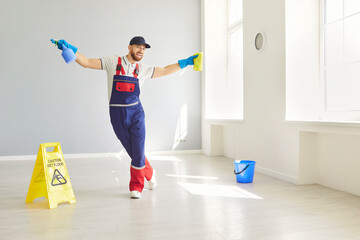 Happy professional janitor entertaining during housework, dancing with rag and pulverizer in bright...