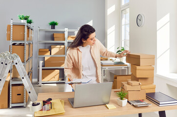 Young confident female employee working in warehouse among cardboard boxes. Woman checking stock and inventory on workplace. Small business owner preparing order for client in storeroom.