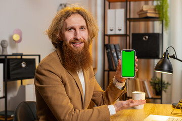 Caucasian bearded businessman holding smartphone with green screen chroma key mock up recommend good application promotional sale offer. Freelancer redhead guy at home office table looking at camera.