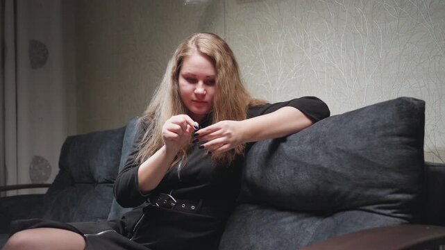Medium view fashion designer in black gown seated on sofa, chatting with smile while guiding thread through needle, attention fixed on hands, living room setting, indoor craft repair moment
