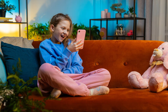 Young child girl texting share messages content on smartphone social media applications online, watching relax movie. Caucasian teenager kid uses mobile phone at home in evening room sitting on sofa
