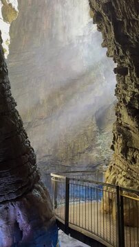 Inside Varone Gorge in Italy, a waterfall descends through dramatic cliffs. Mist swirls in the air, with sunlight beams piercing the canyon, giving the scene a mystical and dreamlike feeling