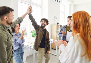 Two men giving high five each other celebrating success, making deal, business achievement or signing contract with group of young people in casual clothes applauding to coworkers in office