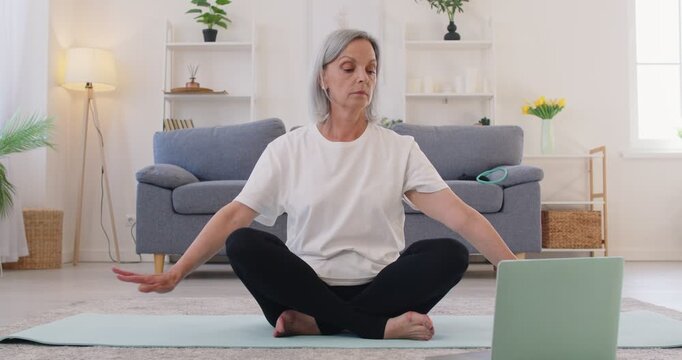 Mature grey haired retired woman sitting in easy pose on yoga mat at home, watching virtual yoga lesson on laptop, and doing arm exercises with online trainer. Wellness, healthy lifestyle concept