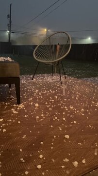 hail hits a patio during a severe hailstorm - ice and hailstones pelt a midwestern backyard. Hail damage.; low angle footage; golf ball size hail striking outdoor furniture, exterior, night.  Vertical
