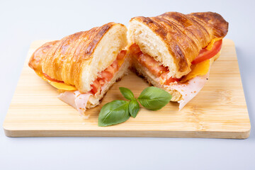 Ham, cheese and tomato croissant sandwich on wooden board with basil garnish. Side angle view of savory pastry, ideal for breakfast, brunch, café menu or lunch concept.