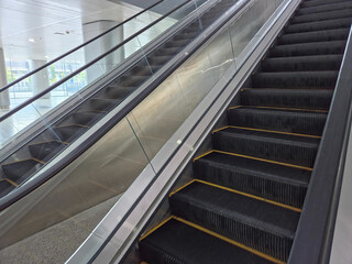 Fototapeta premium Low-angle close-up of modern escalators with black steps and yellow markings, enclosed by glass and metal railings, set in a bright, spacious building interior.