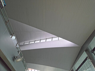 Obraz premium Upward view of a modern interior ceiling with slanted panels, geometric angles, a central window row, and a hanging light, emphasizing clean, dynamic design.