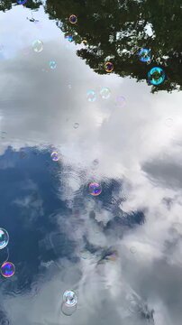 First-person view of blowing iridescent soap bubbles over a lake. Bubbles float and skip on water with sky reflections. Vertical handshot video, dreamy and playful scene.