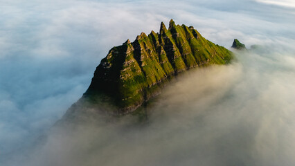 A dramatic mountain rises above a blanket of mist in the Faroe Islands, showcasing rugged cliffs and vibrant greenery.