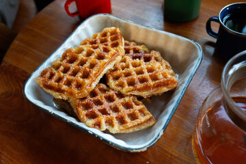 Close-up of freshly baked golden brown croffles served in a ceramic dish on a wooden table. A glass coffee pot and enamel mug complete the cozy breakfast setup.