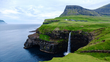 Naklejka premium waterfall tumbles from a green cliff into the Atlantic Ocean, portraying the rugged beauty of the Faroe Islands. Lush vegetation surrounds the area, Mulafossur waterfall near Gasadalur village
