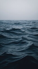 Fototapeta premium Wavy blue water stretches to a hazy horizon under a grey sky. The water reflects the light, creating a dynamic texture