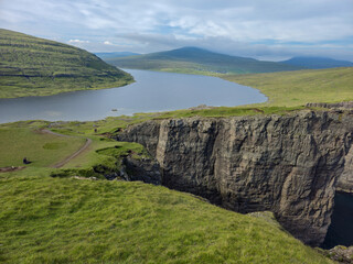 Lush green cliffs rise dramatically above calm waters, with distant mountains framing the tranquil landscape of the Faroe Islands. Perfect for outdoor enthusiasts and nature lovers. Sorvagsvatn lake
