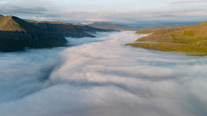 Breathtaking view of soft fog flowing through lush valleys in the Faroe Islands at dawn, creating a magical and ethereal landscape. The sun illuminates the rolling hills, enhancing the serenity.