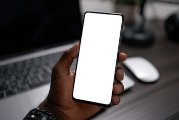 Close-up of a dark-skinned hand holding a blank white smartphone in front of a blurred laptop, mouse, and desk objects on a wooden surface