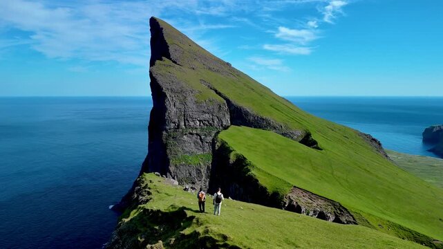 Two hikers stand atop dramatic cliffs in the Faroe Islands, surrounded by lush green landscapes and endless ocean. The blue skies create a perfect backdrop for adventure. Mylingur cliff hiking trail