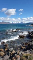 Obraz premium Coastal scene with rocky beach and vibrant turquoise water meeting distant snow-capped mountains under a bright blue sky with fluffy white clouds