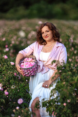 Romantic woman in light dress with curly hair sitting in blooming rose garden with picnic basket, enjoying summer nature outdoors, floral field lifestyle, relaxation and beauty in countryside scenery