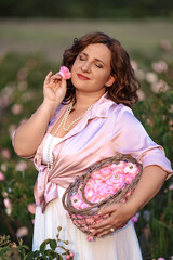 Romantic woman in light dress with curly hair sitting in blooming rose garden with picnic basket, enjoying summer nature outdoors, floral field lifestyle, relaxation and beauty in countryside scenery