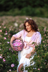 Romantic woman in light dress with curly hair sitting in blooming rose garden with picnic basket, enjoying summer nature outdoors, floral field lifestyle, relaxation and beauty in countryside scenery