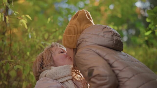 kid kisses mom on lips, playfully pushing mom backward as she smiles, warm autumn light filters through colourful foliage, both in jackets and knit hat, capturing joyful playful family moment