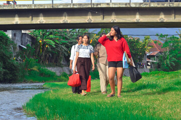A group of girls walk along a riverside meadow trail, carrying supplies and looking around at the scenery, surrounded by shrubs, grasses, and trees on a sunny morning.