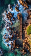 Aerial shot of a wooden boardwalk leading to a rocky ocean shore. Turquoise waves crash against rocks near the coast and the cliffside shows foliage