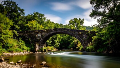 Fototapeta premium A picturesque long exposure view of a stone arch bridge over a serene river, surrounded by lush green trees and under a dynamic, partly cloudy sky.