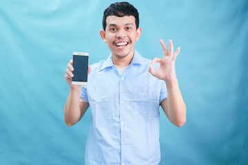 Cheerful Young Man Holding Smartphone with Positive Gesture Against Blue Background