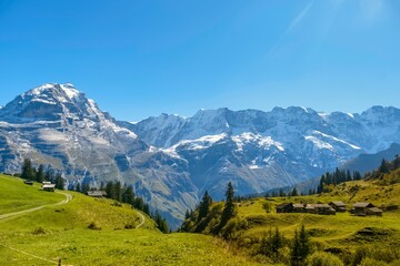 Majestic mountains soar against a bright blue sky, showcasing snow-capped peaks. Lush green meadows and quaint cabins create a serene atmosphere in Switzerland Lauterbrunnen Valley during autumn.