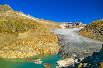 Captivating landscape showcases a massive Rhonegletscher glacier flowing down rocky slopes into a turquoise lake, surrounded by majestic mountains on a sunny day in Switzerland.