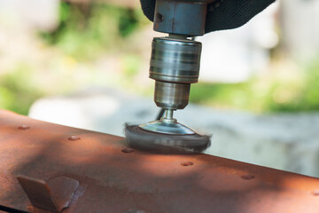 A worker removes rust and corrosion from a metal surface using a wire brush attachment on an electric drill