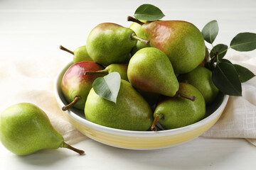 Fresh ripe pears in bowl and green leaves on white wooden table, closeup
