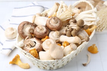 Different raw mushrooms in wicker basket on white tiled table, closeup