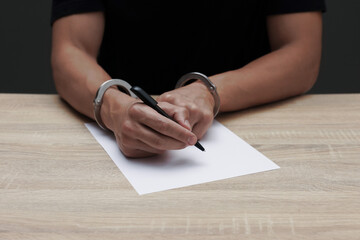 Man in metal handcuffs writing something at wooden table against dark background, closeup