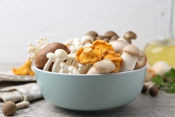 Different raw mushrooms in bowl on grey textured table against light background, closeup
