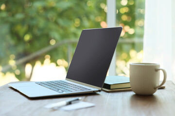 Fototapeta premium Open laptop, stationery and cup on wooden table against blurred green background, closeup