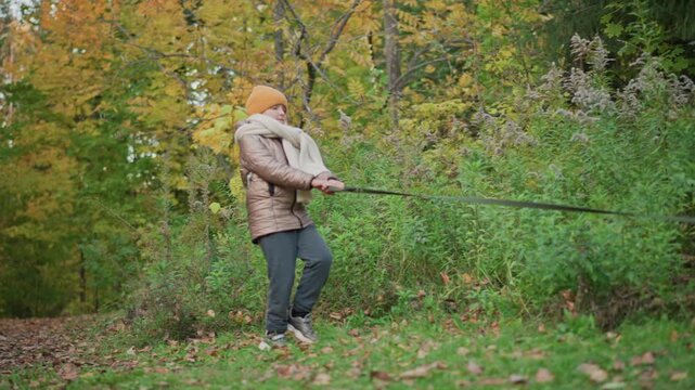 young girl in cozy jacket and knit hat tightly grips leash while being pulled forward by unseen dog or object through vibrant autumn forest trail with scattered leaves and colorful foliage all around