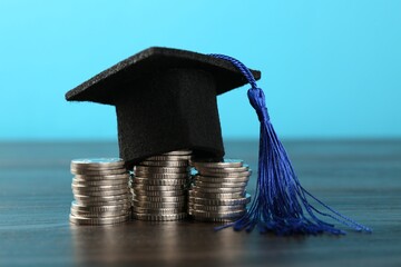 Scholarship. Graduate hat and coins on wooden table against light blue background, closeup