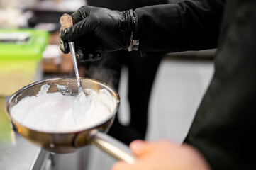 Chef wearing black gloves stirs creamy white sauce in stainless steel saucepan using metal spoon. Food preparation in professional kitchen setting