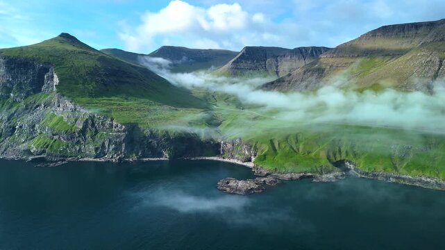 Discover the extraordinary beauty of the Faroe Islands with this aerial perspective. Lush green cliffs rise dramatically from the blue waters, while mist flows gently over the landscape.
