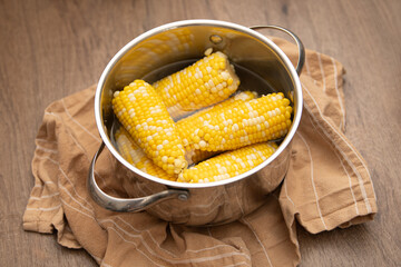 Yellow corn on the cob simmers in a stainless steel pot filled with water. A beige linen towel rests beneath the pot, creating a rustic kitchen atmosphere